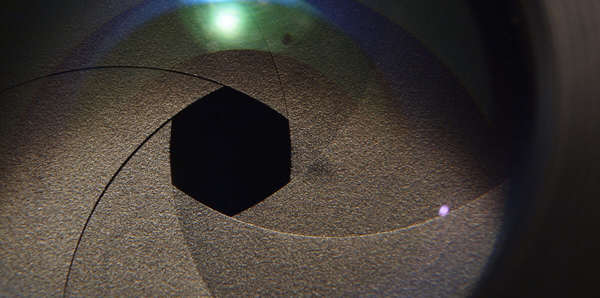 Kameralinse mit Fokus auf eine fast geschlossene Blende, grün-blaue Lichtreflexe in der Linse sichtbar.
